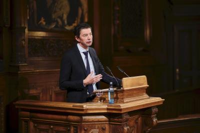 Am holzgeschnitzten Pult der Alten Aula steht ein Mann im dunklen Anzug mit Krawatte, der spricht. Er trägt schwarzgelocktes kurzes Haar.