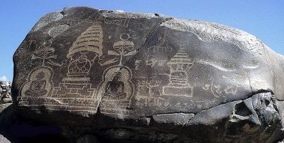 Das Bild zeigt einen großen Fels mit eingeritzten Zeichnungen darauf. Die Zeichnungen zeigen zwei Buddha-Figuren sowie Tempeldarstellungen. Auf den Fels scheint die Sonne, der Himmel im Hintergrund ist blau.