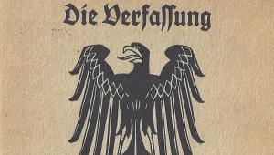 Das Bild zeigt das Deckblatt der Verfassung von Weimar. Auf beigen Untergrund ein schwarzer Adler zu sehen, über dem die Worte "Die Verfassung" stehen. Unterhalb des Adlers steht "des Deutschen Reiches". 