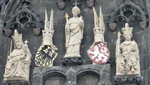 Die Wand einer Kirche oder Kathedrale ist zu sehen. Der dunkelgraue Stein ist verziert mit Applikationen. Im Zentrum des Bildes stehen fünf sandfarbene Figuren, die auf einem Mauervorsprung angebracht sind. Links und rechts sitzen zwei Könige mit Krone, Zepter und Reichsapfel, daneben sind ihre Wappen - ein schwarzer Adler auf gelben Grund und ein weißer Löwe auf rotem Grund - zu sehen, auf denen jeweils ein gesichtsloser Kopf mit Krone prangt. In der Mitte steht eine Figur mit Zepter und Reichsapfel.