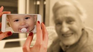 Im Vordergrund sind zwei Hände zu sehen, die ein Smartphone halten. Darauf ist das Bild eines Kleinkinds mit Schnuller zu erkennen. Die Kamera des Smartphones ist auf eine Person im Hintergrund gerichtet, die allerdings nicht mit dem Kleinkind auf dem Bildschirm identisch ist, sondern eine alte Frau mit weißem Haar, die in die Kamera lächelt.